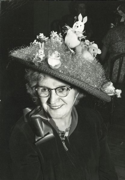 Slightly elevated view of a smiling woman wearing a large hat decorated with artificial Easter bunnies, a live duck, and artificial Easter basket grass.