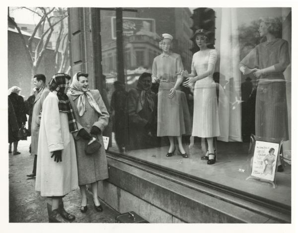Two women stand on the sidewalk laughing and looking at mannequins in a shop window. A caption on the back of the photograph identifies it as from a "teacher's convention."