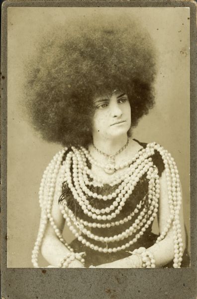 Woman coiffed as a "moss-haired girl" or a "Circassian beauty." A literary tradition exists for the Circassian beauties, but in the United States they achieved popularity as a sideshow attraction promoted by P.T. Barnum, beginning in 1865. The Circassian beauties were said to be especially prized as concubines during the Middle Ages; at Barnum's time they were not necessarily women from the Northern Caucusus region but rather were actresses who wore the distinctive hairstyle Barnum promoted as a trademark of Circassian beauties, hence their other title as "moss-haired girls." The woman is also wearing what appears to be a tight-laced or corseted dress and a multi-strand rope pearl necklace.