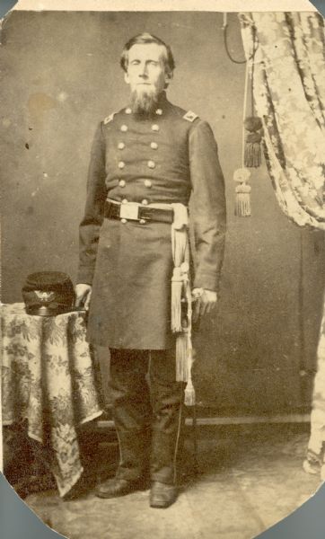 Full-length studio portrait of Colonel George Bryant of the 12th Regiment, Wisconsin Volunteers. His hat is on a table next to him on the left.