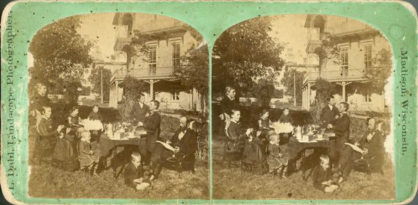 Stereograph of unidentified family posing around two tables in a yard. Includes mother, father, and seven children. In the background is a large, three-story building. Location is not identified.