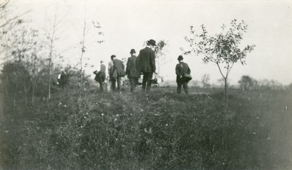 A small group of men, some of them carrying bags, walk in a field among effigy mounds.