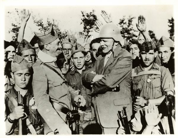 Hans V. Kaltenborn, wearing a helmet, interviews one of Generalissimo Franco's commanders during the siege of Madrid. They are surrounded by young soldiers.