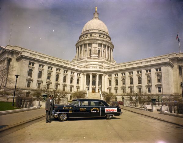 Bob Bender, wearing a business suit and a chauffeur hat, is posing next to "Nellie," his shiny, black 1956 Cadillac Fleetwood parked in front of the Wisconsin State Capitol. He typically parked his cab at a taxi stand in front of the Park Inn. His taxi service, "R.L. Bender Deluxe Cab," flat rates, and contact phone number are in yellow and white letters on the doors and front quarter panel. The white sign on the rear quarter panel commemorates his limousine cab's mileage turning the million mile mark. It reads: "This 1956 Cadillac Has Gone - 1,000,000 - One Million Miles." 
He and Nellie reportedly racked up 250 miles a day on average since purchasing the car used in 1957; at that time, the Special Sixty Cadillac model had 40,000 miles on it. Bender ran passengers between the Dane County Regional Airport and Madison downtown.