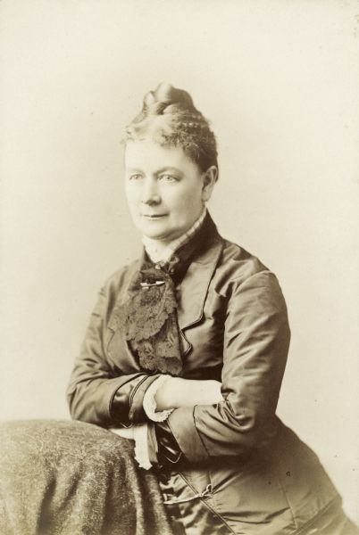 Waist-up portrait of Emma Bascom. She is standing and posing with her arms folded on a cloth-covered table or chair, and is wearing a collar pin shaped like an arrow.