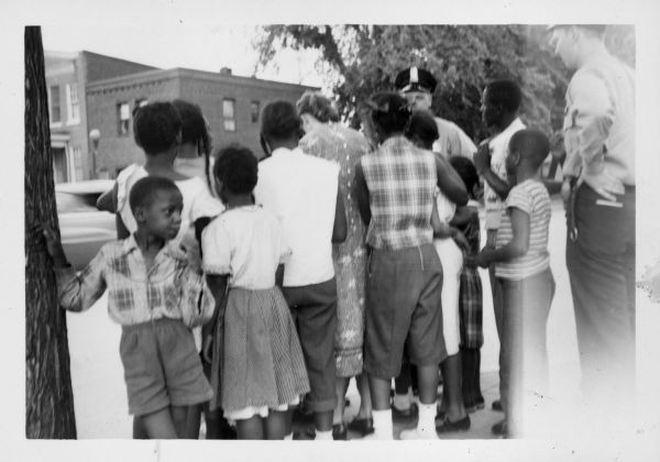 A group of African American children are standing outside the Rosedale Playground. Most of the children have their backs to the camera, including a woman standing in the center. One young boy is standing on the left leaning against a tree and looking to the right. Standing with the group are two white police officers.