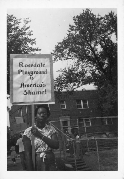 A woman holds a sign that reads, "Rosedale Playground is America's Shame!" as she stands next to the playground. She is protesting the exclusion of African Americans from the playground.