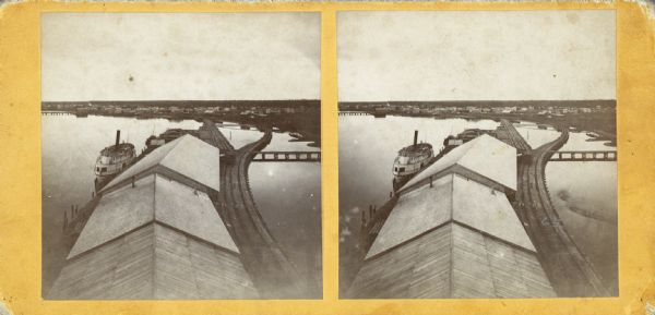 Stereograph of an elevated view of Howard Side of Green Bay. View looks over roofs of long, industrial buildings near railroad tracks that run over the water and curve towards the shoreline on the right. A boat is moored next to the wharf on the left.