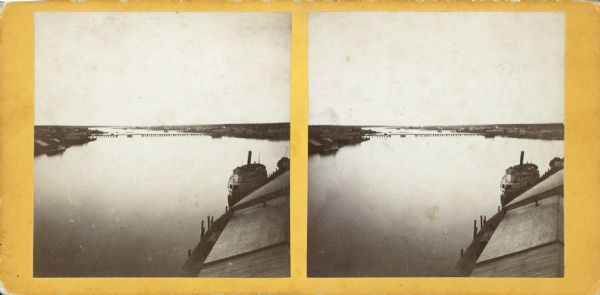 Stereograph of elevated view up the Fox River at Green Bay. View looks over roofs of long, industrial buildings. A boat is moored next to the wharf on the right. There is a bridge crossing the river in the distance.