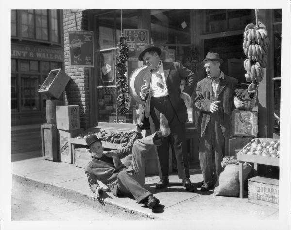 Stan Laurel sitting on the sidewalk with one leg in the air. He is holding on to Oliver Hardy's coat. Hardy is holding a banana and is looking down at Laurel. Another man stands next to Hardy looking down at Laurel. They are standing outside a food market. The still is from the 1927 film "Battle of the Century."