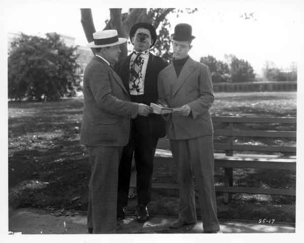 Stan Laurel is signing an insurance policy held by the insurance agent (Eugene Pallette) while Oliver Hardy is looking on. Hardy has ink splashed on his face and shirt. They are standing outdoors in a park. The still is from the 1927 film "Battle of the Century."