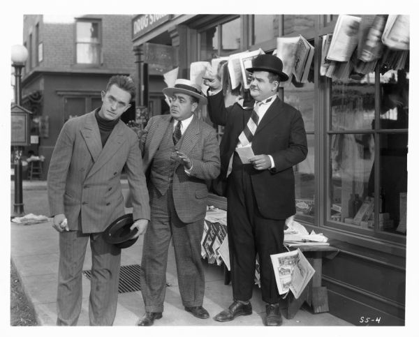Oliver Hardy is pointing to Stan Laurel while the insurance agent (Eugene Pallette) looks at Laurel with concern. Laurel is holding his hat in his hand and has a blank look on his face. They are standing on a sidewalk. The still is from the 1927 film "Battle of the Century."