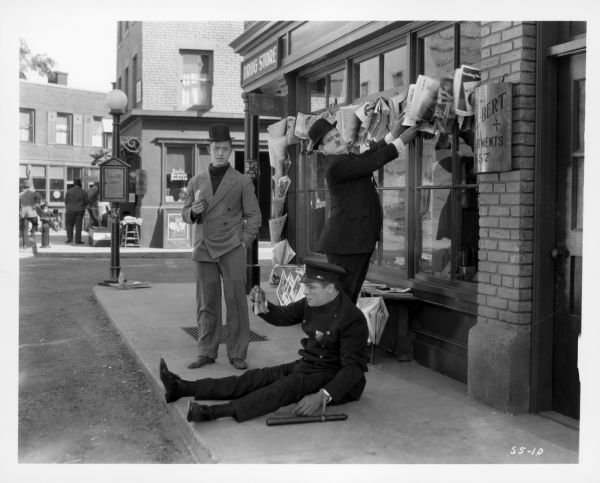 Stan Laurel and Oliver Hardy standing on a sidewalk. They are both looking at a police officer sitting on the sidewalk holding a banana peel. Laurel is holding a banana. The still is from the 1927 film "Battle of the Century."