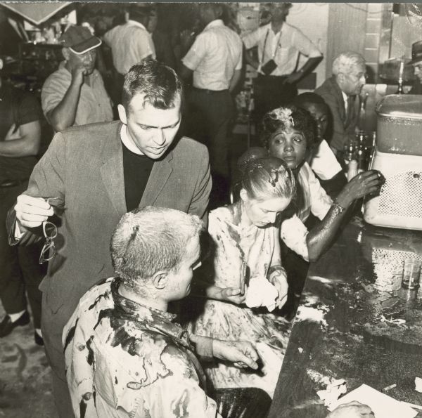 A slightly elevated view of a civil rights sit-in at a Woolworth's lunch counter. John Salter is seated in the foreground. Sitting next to him is Joan Trumpauer, and Anne Moody is sitting next to her. Tom Beard is seated next to Moody. People have poured ketchup, sugar and various liquids on them. Reverend Ed (Edwin) King is standing with them. He is speaking to Salter and is holding Trumpauer's eyeglasses.