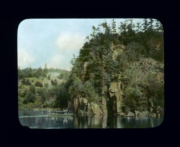 View across water towards people in an excursion boat on a river passing rocky and hilly shoreline covered with trees and plants. There is a man in another boat near a cave in the rocks at water level on the right.