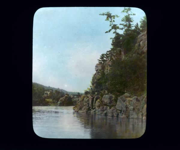 View across water with rocky bluffs rising from the river on the right. A rock formation on the bluff resembles a human profile. In the distance on the left is a large building or house surrounded by trees.