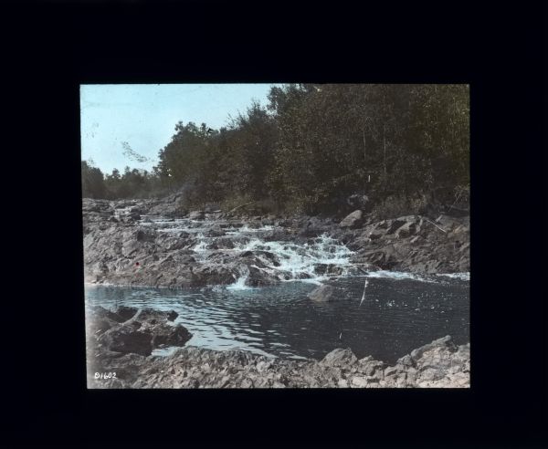 View from rocky shoreline across a pool in a river with a small waterfall in the background. Trees and shrubs line the opposite shoreline.