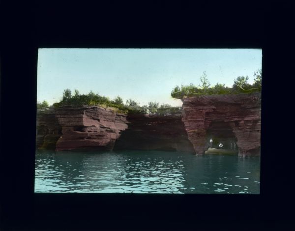 View across a river towards rock formations on the far shoreline. Several people are standing in a boat or canoe in a cave on the right.