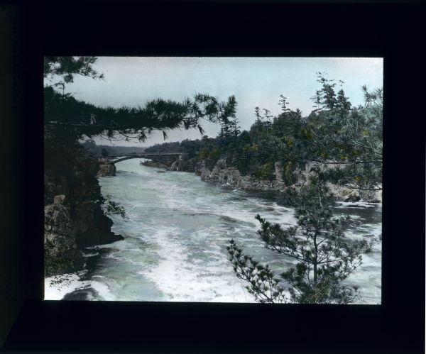 Elevated view through branches of pine trees of a rushing river and a bridge in the distance spanning the river from rock cliffs.