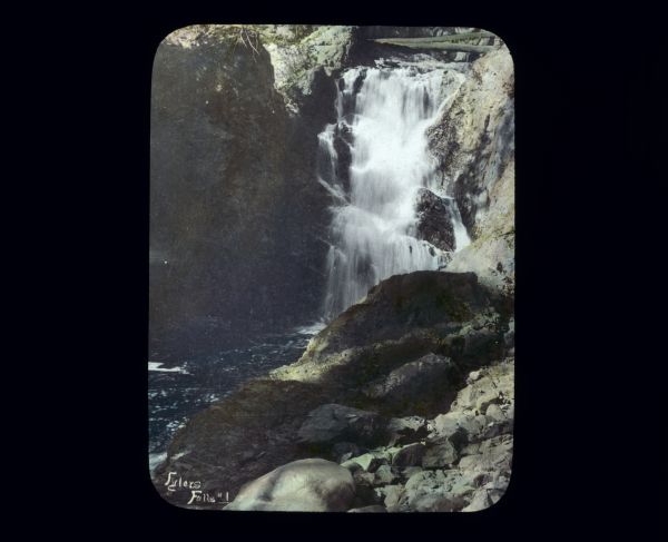 View from rocky shoreline looking up at Fylers Falls, a waterfall surrounded by rock cliffs.