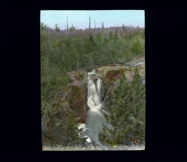 Elevated view looking down at a steep waterfall surrounded by cliffs. The tops of the cliffs and surrounding area are covered by trees and shrubs. A large tree trunk has fallen into the pool and is leaning against the rocky cliff.