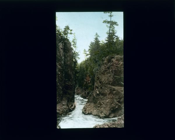 View looking down river as it is flowing through narrows between two steep cliffs. Trees and plants are growing on top of the cliffs.