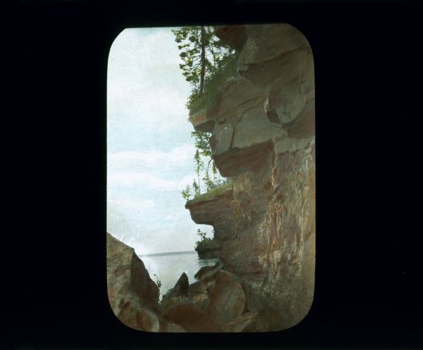View through large rocks towards a lake. On the right cliffs with overhangs topped with trees run along the shoreline. A tree-lined shoreline is in the far background.