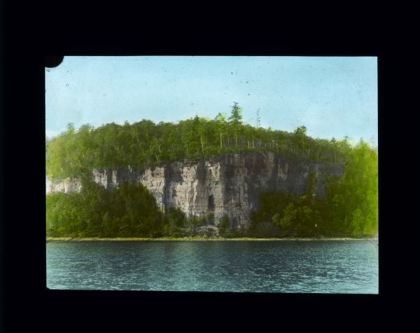 View across lake towards steep bluffs. Trees are growing along the shoreline and on the top of the bluffs. At the base of the cliffs in the center is a scaffolding leading up to a large opening, perhaps a cave, in the face of the cliff.