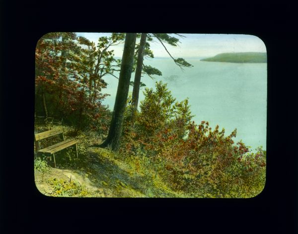 A wooden bench at the top of a slope overlooking a lake. A wooded shoreline is in the far distance.