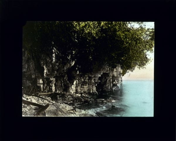 View along shoreline of a lake towards a man standing near the mouth of a cave at the waterline. The cave is in steep, eroded cliffs, and trees and shrubs are hanging over the edge of the cliffs above the cave.