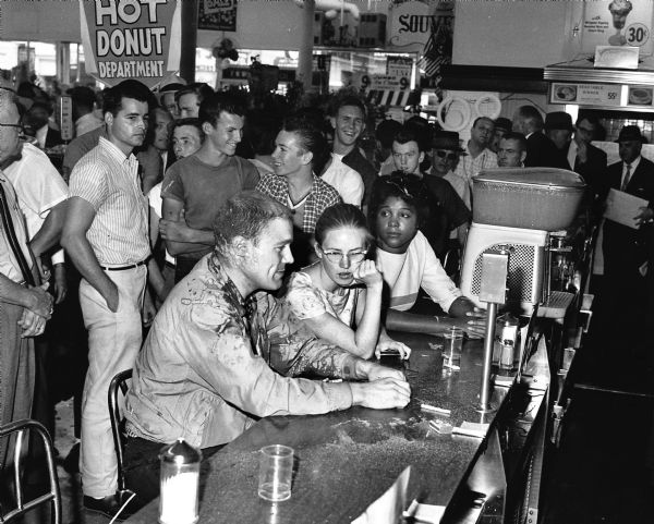 Three civil rights activists sit at a Woolworth's lunch counter in a non-violent demonstration. From left to right are Professor John Salter, Joan Trumpauer, and Anne Moody. A group of laughing, white teenage boys stand behind them. They have poured sugar, mustard, and ketchup on the protesters.