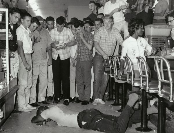 Civil rights activist Walter Williams lying face down on the floor near the lunch counter at Woolworths, where he was taking part in a sit-in. He had just been struck by a glass object. A crowd of white teenage boys are looking down at him after having poured drinks on Williams. Pearlena Lewis is sitting at the counter and UPI reporter Cliff Sessions is speaking to her over the counter.