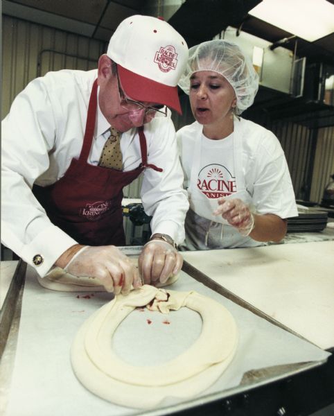 Wisconsin Governor Tommy Thompson folding a kringle. This image appeared in <i>The Chippewa Herald</i> on August 31, 1999. The caption read: "Darleen Tennessen, right, of Mount Pleasant, gives some pointers to Wis. Gov. Tommy Thompson on how to properly shape a kringle, during a tour of Racine Danish Kringles, Monday, in Racine." Kringle is a Danish pastry particularly associated with many Racine bakeries.
