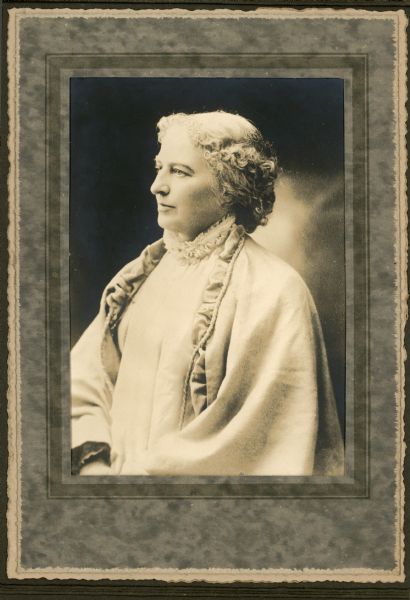 Waist-up studio portrait of Emma Dryer. She is wearing a light-colored cloak, or jacket with large sleeves, over a ruffled, high-necked blouse.