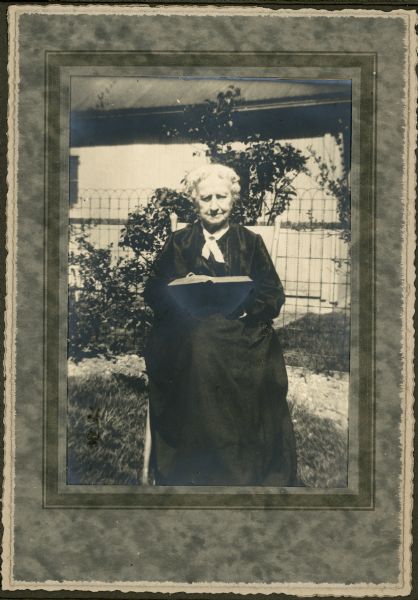 Candid portrait of Emma Dryer, seated outdoors. She is wearing a dark-colored dress and holding a book. There is a fence and foliage behind her.