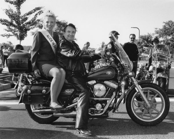 Alice in Dairyland, Courtney Ott, wearing a crown and a sash, is sitting on the back of a motorcycle driven by Governor Tommy Thompson at the start of the annual Harley-Davidson rally.