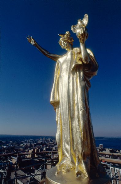 Close-up of the statue "Wisconsin," created by Daniel Chester French and situated on top of the dome of the Wisconsin State Capitol, immediately after cleaning and regilding. This project was part of the restoration of the state Capitol building, overseen by Tony Rajer. Scaffolding surrounds the statue. Part of the Madison skyline and the shoreline of Lake Mendota is below in the far background. 
