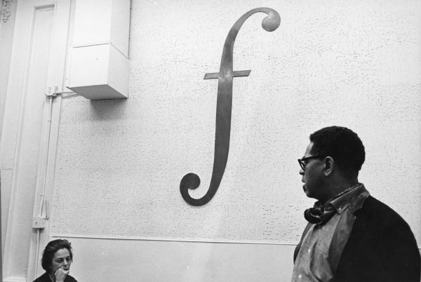 Dizzy Gillespie looking at Shirley Clarke, sitting on the left with a contemplative look on her face, during a recording session for the film "The Cool World." They are in a recording studio and there is a large letter "f," or "forte" which means loud in music, on the wall.