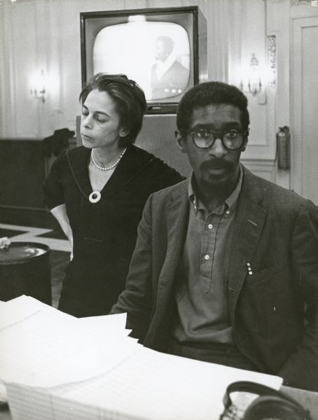 Shirley Clarke standing next to Mal Waldron, who is sitting. There is a television monitor behind them. They are working on the film "The Cool World."
