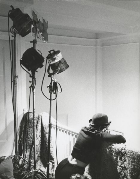 Shirley Clarke is seen kneeling against a stair banister during the shooting of the film "The Cool World." She is wearing a derby hat.  There are three lights on stands to her left, and rugs are draped over the banister.