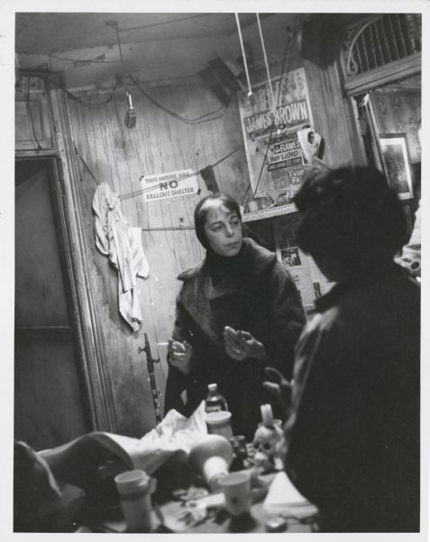 Shirley Clarke standing on the set of the film "The Cool World." She is standing in a rundown house or apartment and is wearing a scarf and coat and holding a cigarette. A woman stands with her back to the camera in the foreground. A James Brown poster, and a sign that reads: "This House Has No Fallout Shelter," are on the wall in the background. There appears to be a microphone handing from the ceiling.