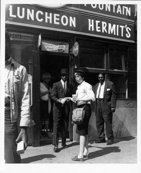 Shirley Clarke standing on the sidewalk outside of a restaurant while shooting the film "The Cool World." She is holding a glass and is being handed something by a man while she is looking towards someone else.