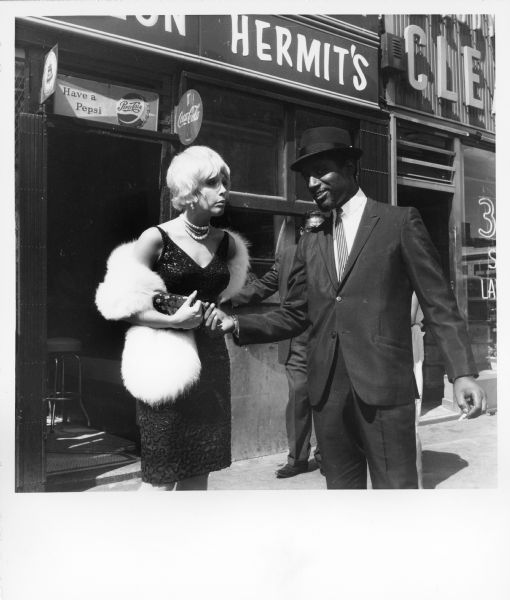 An actress and actor in the film "The Cool World" are standing on the sidewalk outside a restaurant. She is wearing a cocktail dress and a fur wrap. He is dressed in a suit and wears a hat.