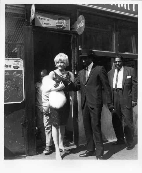 An actress and actor standing just outside the entrance of a restaurant while shooting the film "The Cool World." The woman is wearing a cocktail dress and a fur wrap. The man is wearing a suit and a hat.  Another man in a suit is standing behind the man on the right. Just inside the door is a boy standing directly behind the actress.