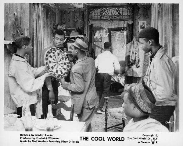 A publicity still from the film "The Cool World" showing six African American teenage boys in a rundown house or apartment. Two of the boys are holding a dart board while three others are watching. Another boy is walking away in the background.