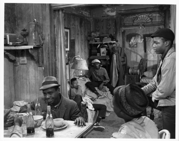 Still photograph from the film "The Cool World" showing five African American teenage boys in a rundown apartment or house. Two boys are sitting on a couch in the background, while two others are sitting at a table. Another young man is standing on the right. There are dishes on the table and three bottles of Pepsi.
