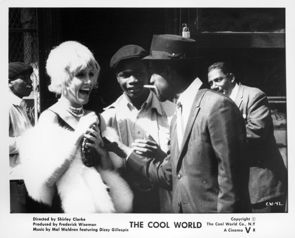 Publicity still from the film "The Cool World" showing a woman wearing a cocktail dress and fur wrap laughing while looking at a man dressed in a suit and smoking a cigarette. A young man is standing behind and in between them and looking at the man. Two other men are standing in the background.
