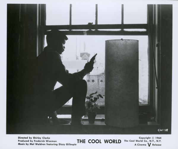 A publicity still from the film "The Cool World" showing an African American teenage boy standing next to a large window holding a gun.