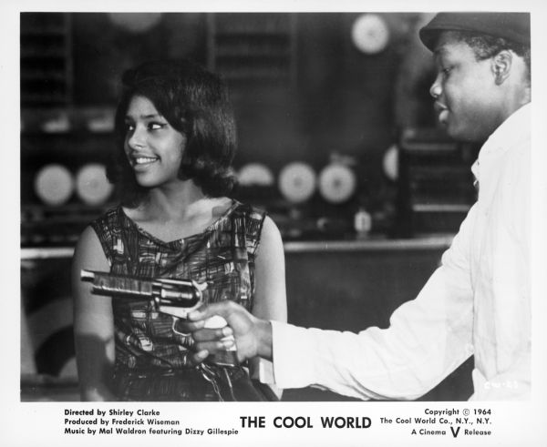 A publicity still from the film "The Cool World" showing an African American teenage boy holding and pointing a gun. An African American teenage girl is standing behind the gun and is smiling while looking towards someone off-screen.