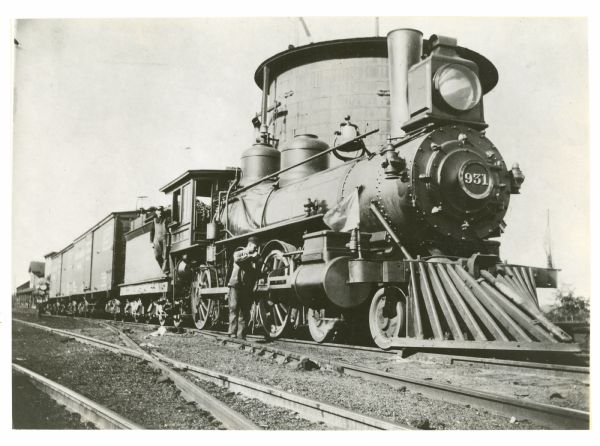 View down railroad tracks towards engineer Louis Peterson standing in the gangway next to Engine No. 931. Another man is standing on the tracks near one of the locomotive's wheels. A large water tower is behind the engine.